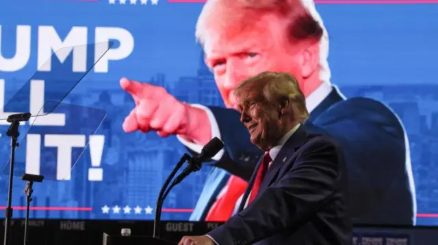 Trump stands at a podium in front of a large photo of himself in the background.