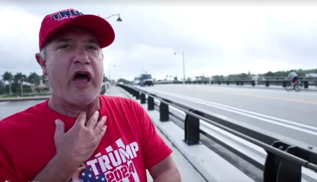 Rich speaking whilst holding one hand to his throat and standing next to a bridge. he wears a Trump hat and t-shirt