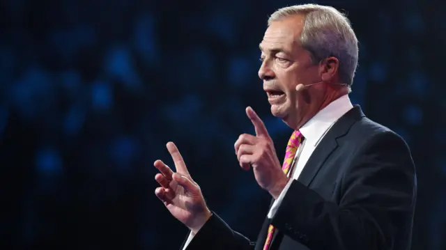 Nigel Farage speaking on a stage. He is wearing a suit with a patterned yellow and pink tie.