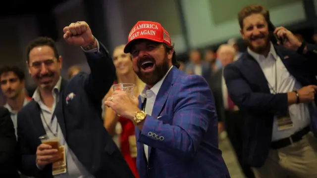 A man wearing a Maga cap and drinking a beverage smiles