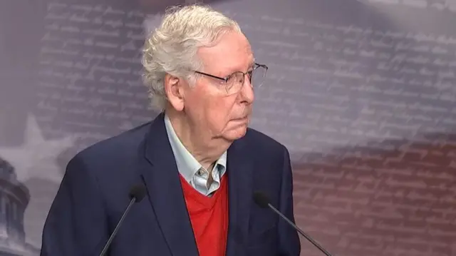 McConnel standing in front of two microphones with his head turned slightly to his left. He wears a red jumper, blue shirt and navy suit jacket and glasses.