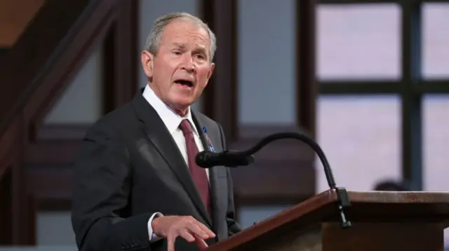 George W Bush, wearing a black suit with red tie, speaks at a podium