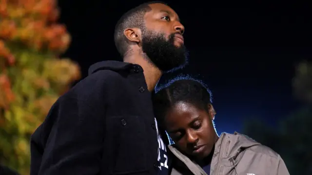 A woman checks her phone as she leans against a man, both of whom look pensive