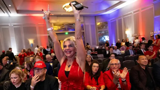 A woman, wearing a red top raises her hands in the air at a Trump watch party