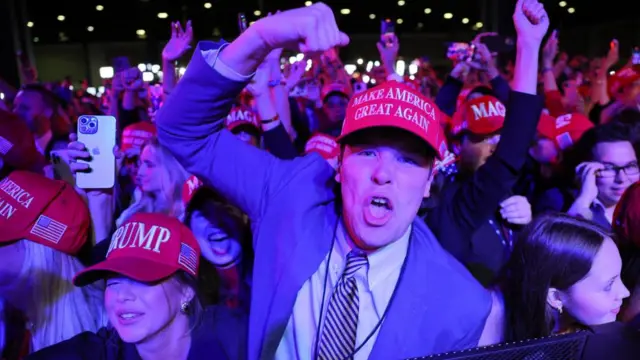 Ecstatic Trump supporters cheer wearing red MAGA hats, with a young man in the centre with his arm raised