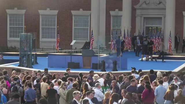 United States flags line the walkway to the front of a stage carpeted in blue. Bullet proof screens shield the lectern.