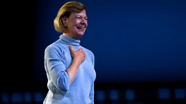 Tammy Baldwin on a stage in a light blue turtleneck jumper. She has short, dark blonde hair, and is holding her hand on her chest while smiling.