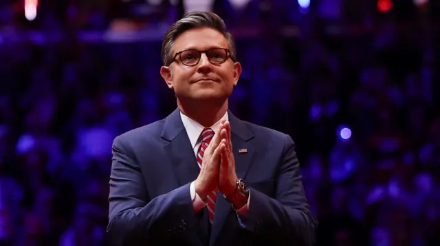 Mike Johnson holding his hands together. He is wearing a blue suit with a red and white striped tie. A crowd of people sitting in the audience of a rally can be seen blurred behind him