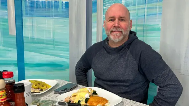 Monty Najar, a bald man wearing a jumper, sits at a table with an egg breakfast in front of him