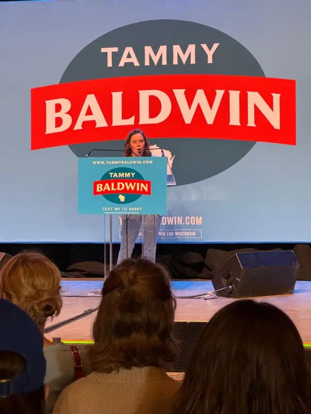 Madison Lieutenant Governor Sara Rodriguez speaks into a microphone in front of a large banner saying Tammy Baldwin