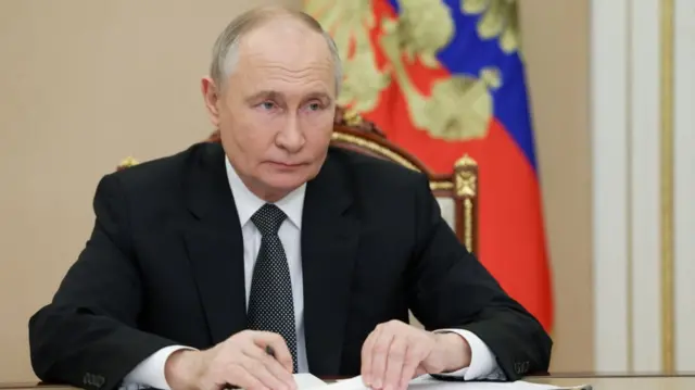 Vladimir Putin sitting at a desk touching pieces of paper while holding a pen in his right hand. He's wearing a black suit, white shirt and patterned black ties while staring ahead