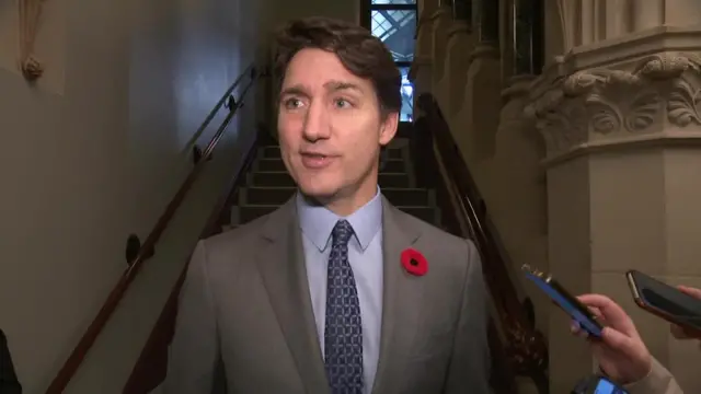 Trudeau speaking to reporters in front of a stair case. He wears a grey suit and a poppy