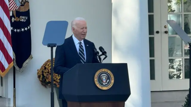 Joe Biden in blue suit and striped blue tie with white shirt speaks behind a lectern in Rose Garden behind the West Wing of the White House