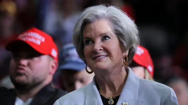 Susie Wiles mid-shot from shoulders up. She's in a white suit jacket and dark shirt. She has short white and grey hair and is wearing golden hoops earrings