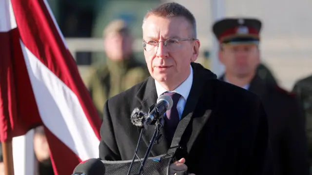 Latvia's President Edgars Rinkēvičs stood at a podium with a microphone. He is wearing a suit with a black coat over it. He has very short, dark blonde hair and thin-rimmed glasses