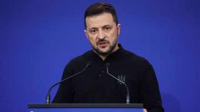 Volodymyr Zelensky stood at a podium in front of a plain dark blue background on a stage. He is wearing a black jumper.