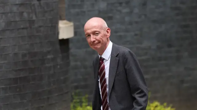 McFadden in grey suit and burgudy, grey and ivory stripes tie, looks at the camera as he walks toward Number 10