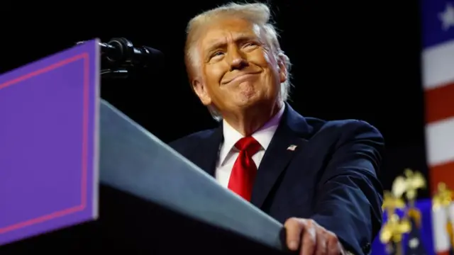 Donald Trump delivers speech looking content as he leans on a podium. He's wearing a dark blue suit, white shirt and red tie.