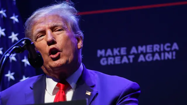 Donald Trump in a navy suit with a red tie stood at a podium in front of a microphone, speaking. He is stood in front of a backdrop that says Make America Great Again