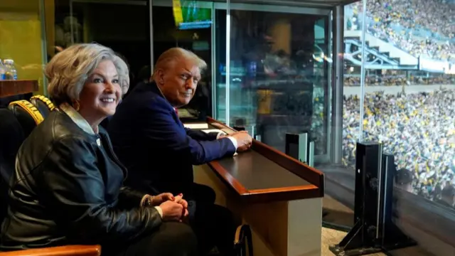 Donald Trump and Susie Wiles sat in a box watching football. Trump is in dark blue suit with red tie, his head turned to the right. Wiles is in a white blouse and leather jacket also looking right. Football fans sitting outside in the stadium visible to the right