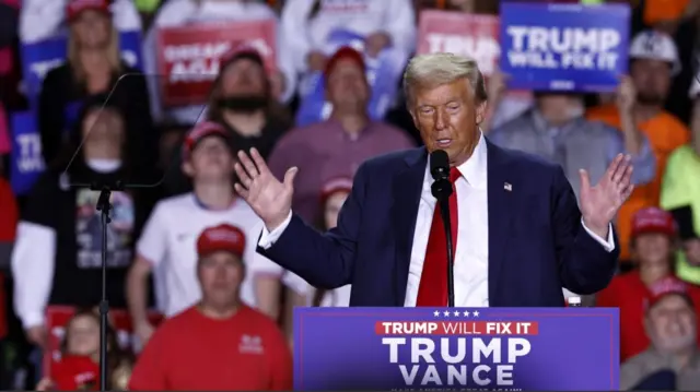 Donald Trump speaks on stage. He's wearing a dark blue suit, white shirt and red tie, and has a US flag pin on his left lapel. Both his hands are raised to his side. Blurred background of supporters holding pro-Trump signs