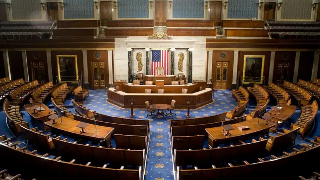 Inside view of empty House of Representatives chamber. Blue patterned carpet all over the floor, seating is made up of long wooden benches with backrest