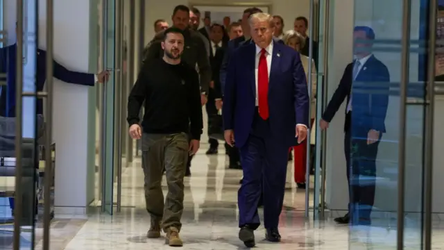 Volodymyr Zelensky in green military trousers and black long-sleeved shirt walks next to Donald Trump in dark blue suit, white shirt and red tie. Behind them are members of their entourage and two men holding doors open