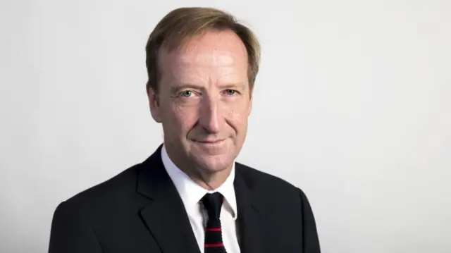 Headshot of Alex Younger against a white wall wearing a black and red tie and a black suit jacket with a white shirt