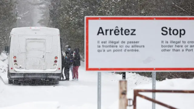 A sign by a snowy road warns against illegal crossing in English and French. Behind it, people pile into a white van