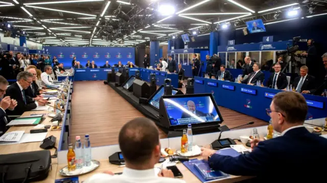 leaders of the EU 27 meet with other European leaders in Budapest. The group are sitting next to each other at desks forming a rectangular with camera placed in the walkable centre