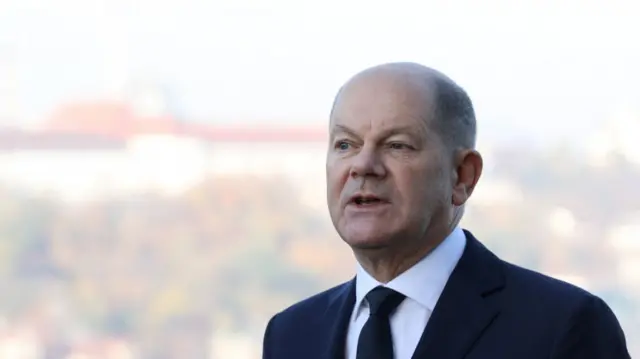 German chancellor OIaf Scholz delivers speech outdoors. He's in black suit, black tie and white shirt. Mid-shot cuts off just below the shoulders, in background is blurred trees and building