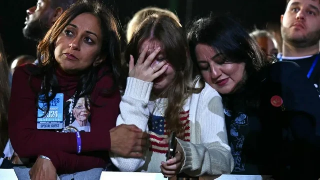 A woman on the right leans her head on the shoulder of another, who covered her face with her hand, crying. Another woman on the left looks despondent