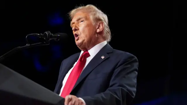 Donald Trump delivers speech behind lectern. Image cuts off just below the mid-riff. He's in a black suit, white shirt and red tie, his left hand grips the side of the lectern