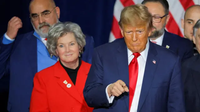 Susie Wiles stands next to Donald Trump, who is flanked by other aides