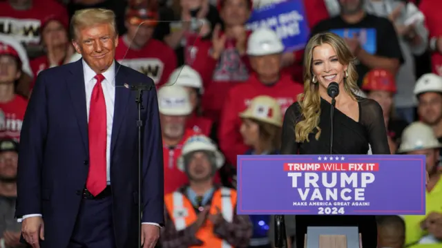 Donald Trump standing behind Megyn Kelly, who is speaking at a microphone on top of a lectern which reads 'Trump/Vance 2024'