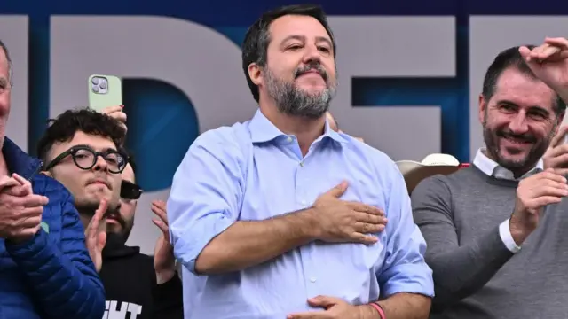 Matteo Salvini reacts during a traditional Lega party rally in Pontida. He's wearing a light blue shirt and is holding his right hand over his heart. Next to him are other men clapping and cheering.