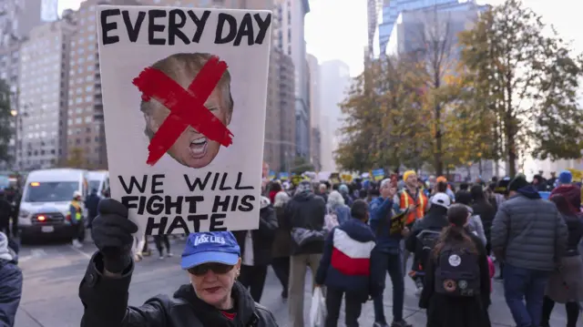 A woman in a crowd of protesters holds up a sign with a picture of Donald Trump's face with a red cross over it alongside the words Everyday we will fight his hate