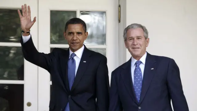 Barack Obama and George Bush, both wearing darks suits, patterned blue ties and US flag pins on their jackets's left lapel, walk to the Oval Office together