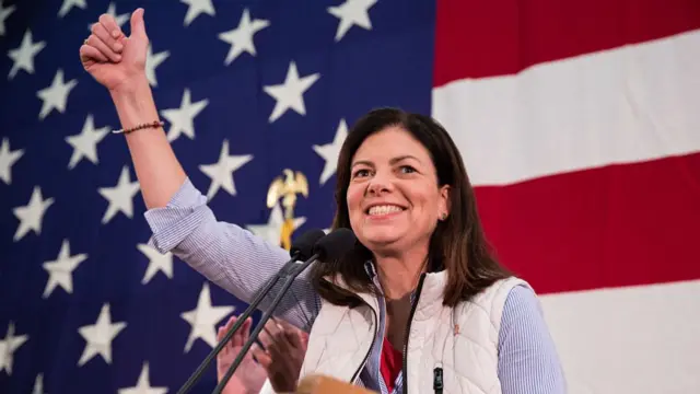 Kelly Ayotte waves at a podium with an American flag in the background