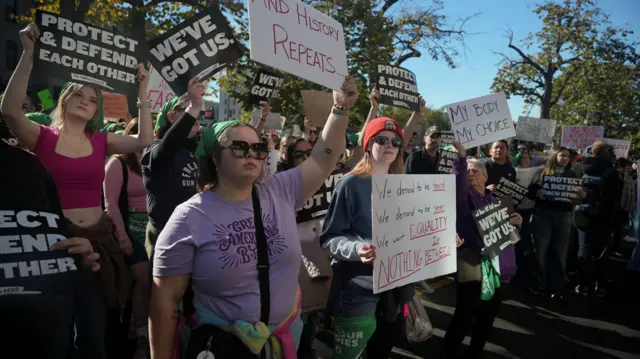 Women stand holding placards in a crowd. One says And History Repeats. Another sign says Protect & Defend Each Other. A third says We've Got Us, and another says My Body My Choice