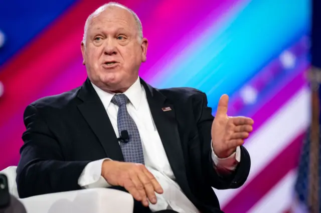 Tom Homan in front of a purple background speaking to a crowd