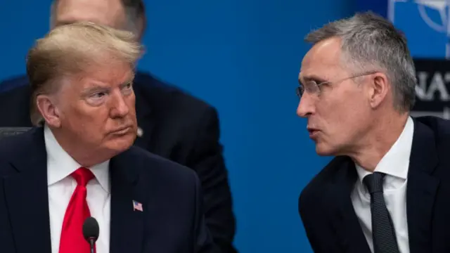 Donald Trump (L) mid shot. He's in dark suit with white shirt and red tie, US flag pin on left lapel and mic in front of him. Jens Stoltenberg (R) leans sideways to speak to Trump. He's in a black suit, white shirt and black tie. Behind them a man covered by Trump and a blue background with Nato logo on top right