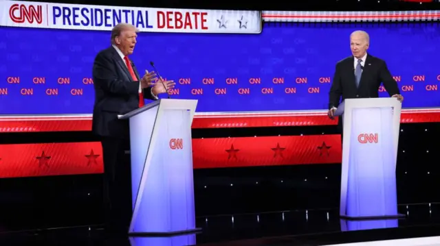 Biden and Trump stood at podiums in front of a blue and red background with the word CNN Presidential Debate written on it. Both are wearing dark suits and white shirts, Trump has a red tie and Biden a patterned blue one