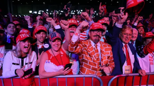 Group of Trump supporters celebrate outside. In first row, left to right, are a blonde woman with Trump red cap; a woman in a red rank top with a US flag cap; a middle-aged man with a suit in a brick wall pattern and a MAGA hat; a middle-aged man with his right hand raised in dark blue suit and red tie