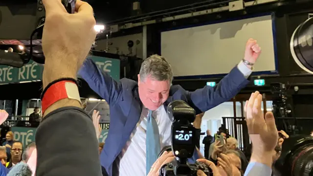 Paschal Donohoe celebrates his win in a navy suit and teal tie, surrounding him are press photographers.