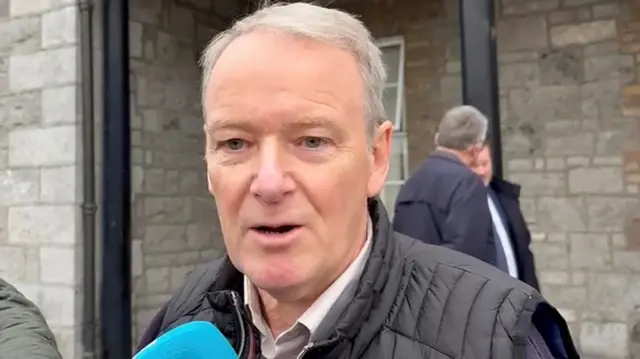 Paul Stanley wears a black Gillet with a white shirt underneath. He is speaking into a blue microphone, standing in front of a grey brick building.