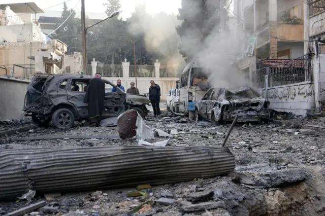 Burnt out cars sit on a street, one still smoking, as people inspect damage