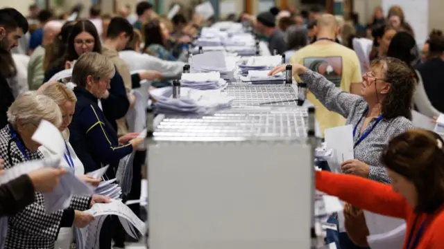 Several women stand either side of a row of grey ballot boxes, counting the votes.