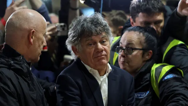 Gerry Hutch wears a navy suit and white shirt, he looks disgruntled. Behind him are security officers in black coats and high-viz jackets