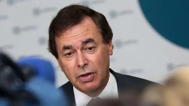 Alan Shatter wears a black suit with a white shirt, behind him is a white background